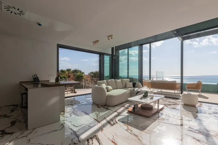 Open living room of THE ONE in Higuerón, with a curved cream sofa on a marble floor and glass walls looking out to the sea
